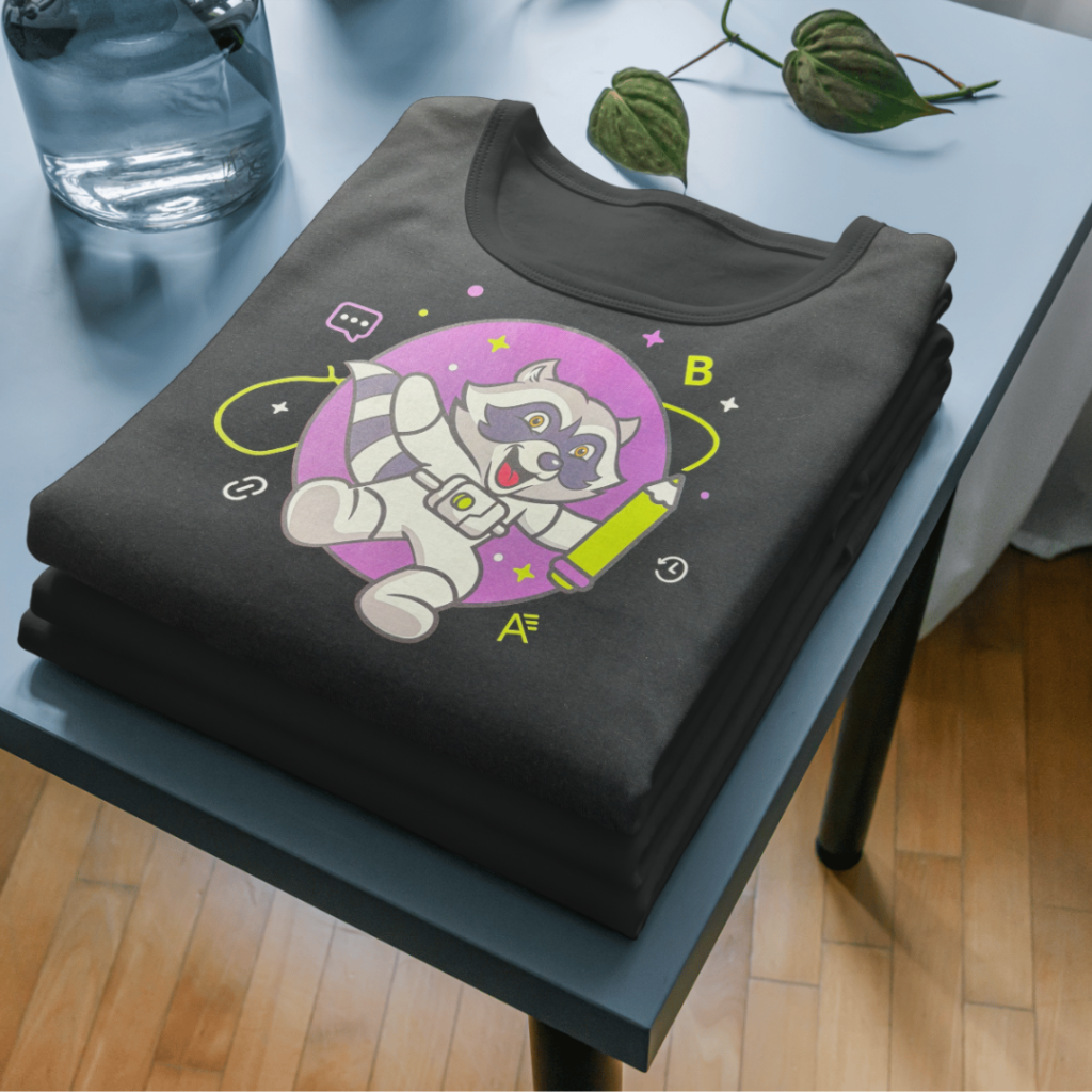A stack of folded dark gray t-shirts is shown, the top one featuring a cartoon raccoon graphic. The raccoon is depicted in a white spacesuit, holding a green pencil, and surrounded by a purple circle with stylized stars and symbols. The letters "A" and "B" are also visible near the raccoon. The t-shirts are neatly stacked on a blue-gray surface, with a glass of water and a plant visible in the blurred background.