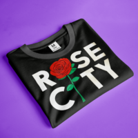 A folded black t-shirt with "ROSE CITY" printed in white letters and a red rose graphic is displayed on a purple background. The word "ROSE" is stacked vertically on the left, with the "O" replaced by the rose image, and "CITY" is stacked vertically on the right. A small tag indicating "M Size 100% Cotton" is visible on the shirt's collar. The shirt is neatly folded and positioned diagonally on the solid-colored backdrop.